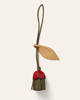 A small leather accessory with a flower strap and leaf-shaped tab, attached to a dangling flower shaped charm in red and natural beige, shown on a plain light beige background.