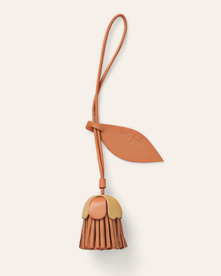 A small leather accessory with a flower strap and leaf-shaped tab, attached to a dangling flower shaped charm in red and natural beige, shown on a plain light warm-gray background.