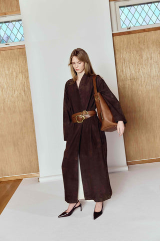 Woman in a brown outfit with a belt and large bag standing in a minimalistic room.