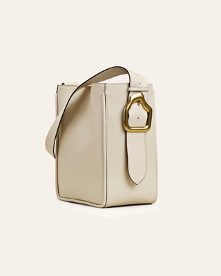 A white leather tote bag with a custom-made brass buckle, an adjustable shoulder strap on beige background