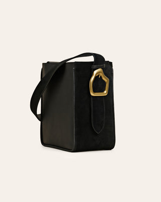 Black suede tote bag with a custom-made brass buckle, an adjustable shoulder strap obn a beige background