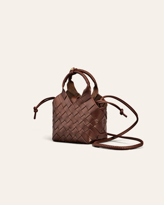 A small brown woven leather handbag with a top handle and a long matching shoulder strap, photographed on a plain light warm-gray background.