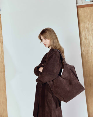 Woman wearing a brown coat with a large bag against a light-colored wall.