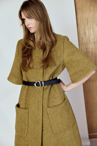 Woman wearing a long mustard yellow coat with a belt against a white background with a leather belt