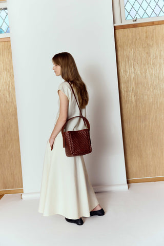 Woman holding a brown woven handbag in a minimalistic setting with wooden panels and a white wall.