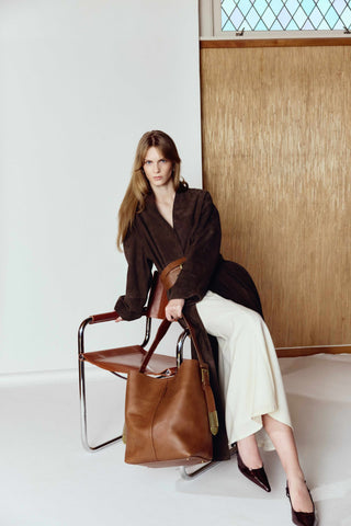 Woman holding a brown leather bag in a minimalistic room with a window and wooden panel.
