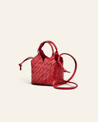 A small red woven leather handbag with top handles and a long shoulder strap, photographed on a clean off-white background.
