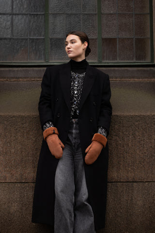 A person wearing brown shearling leather mittens and a black coat with a collar, posing against a window.